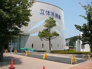 灰色の波板のようなもので覆われた壁を持つ円柱形の建物の側面。濃い青字で立体映画館と書かれてある。