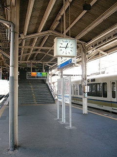 跨線橋階段前の風景。手前に駅名標と時刻表。階段上り口上には黒、緑、青、橙で色分けされた番線案内が吊るされている。