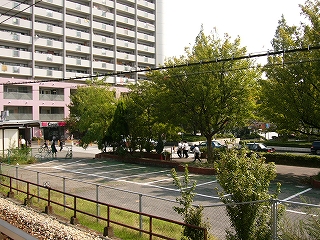 狭い駐車場、そして緑の並木、その向こうにロータリーがある。左奥には高層マンション。