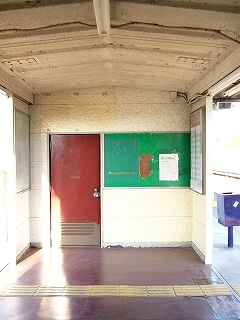 反対側。クリーム色に塗られた天井と壁の駅舎内。緑色の掲示板、とび色の扉。