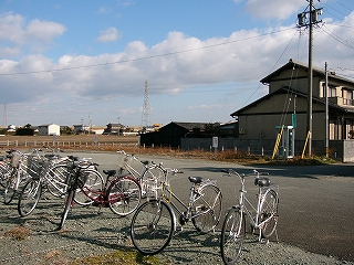 手前に自転車の並び、アスファルトの敷地を挟んで、人家が一件写っている。