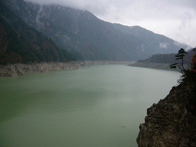 雲がかかり陰鬱な空の下で灰白色の岩肌が均等な幅で縁取りする普通の緑の黒部湖