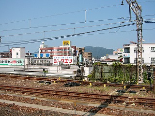 手前は茶色の線路、奥には敦賀市の建物が青空の下に広がる。