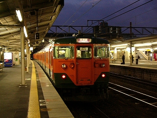 停車中の緑と橙の急行型列車の先頭車。方向幕は紫地に白い字で「草津線」と書かれている。