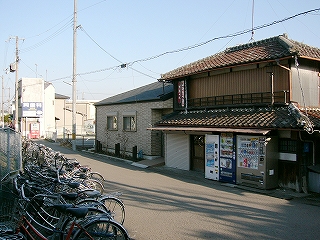 1.5車線程度の道の左側に整然と並べられた自転車、右側には自販機のある古い民家。