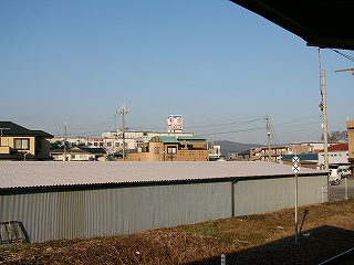 ホームから見える風景。灰色のトタンで覆われた平屋の横にかなり長い倉庫のような建物、その向こうに家々が見える。