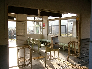 間口にある白い鉄の小さな柵とステンレスのきっぷ入れ。その向こうに窓の多い駅舎内が覗える。