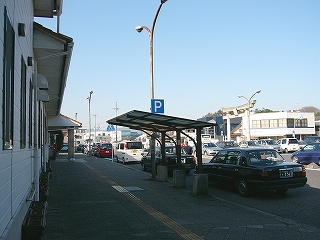 個人宅のガレージの片持ちの屋根が一つ設置された駅舎前の歩道。右手にはタクシーが並んでいる。