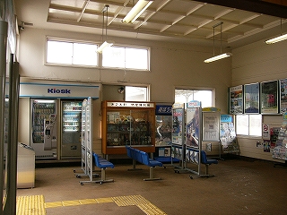 白壁で高い天井の待合室部分。突き当たりの壁に沿って自販機、物産陳列ショーケースが設置され、その前に青色の個人掛けの椅子が並んでいる。