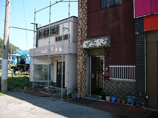 1.5階建ての小さな白い壁の建物。建物前面の上部には「お食事処」と黒字で掲出されてある。建物は足場で囲まれていた。
