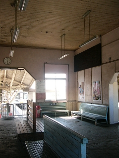 ホームへの間口を突き当たり右に見て駅舎内を俯瞰する。駅舎内中央に両側とも座れる木製のベンチ二脚、右奥の隅に先ほどの水色の木製のベンチが二脚置かれている。