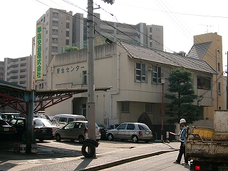 2階建てで白く塗られたコンクリートで造られた、少し薄暗い感じの建物を斜め左に見て。