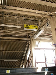 階段を上りきってしまう数段手前から天井を見上げて。天井には横長でコンパクトな黄色に黒い字で書かれた「←北出口 南出口→」の案内板。直進すればまたすぐに階段がホームへ下りる。