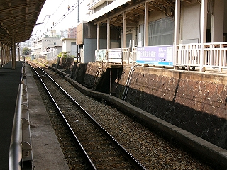 左にはレールに面するように立てられたステンレスの柵、中央に日陰の1線の線路、そして右手にはホーム上の右手には焦げ茶に変色した石垣の上にある駅舎の回廊。