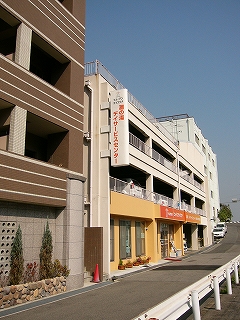 左に3階建ての新しい建物。上二階は白色で駐車場、一階はオレンジ色を基調としたお店で明るい感じ。青空の下で映える。
