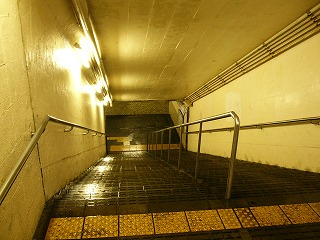 電灯に照らされた階段のトンネル内部。トンネルの壁はクリーム色。コンクリートの階段は濡れている。