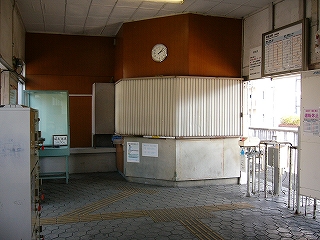 壁を斜めに切って造られた出札口と改札口を駅舎の中の端から見て。出札口の左脇には何も入っていない大きいガラスケースと書き物をする台が置かれている。壁は茶色、天井は白色。