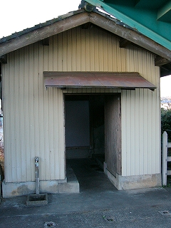 白い波板が壁に貼られた、妻面に入り口のあるトイレ。屋根は瓦葺で、作りは木造。