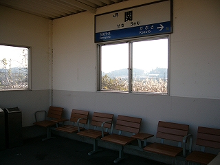 待合室の中。こじんまりした簡単な待合所で、中には二箇所窓があり、明るい茶色の一人掛け用の椅子が並べられている。天上付近の壁には駅名表もあった。