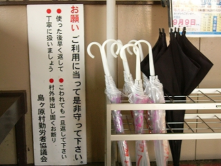 直方体の白い網の傘立てに4,5本刺さったビニール傘。左には白地に黒い字で縦書きで書かれた、縦長の説明板