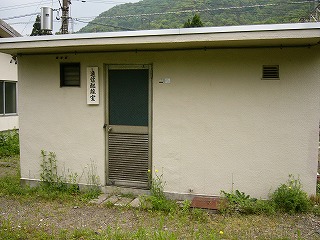 白く四角く小さい平屋の建物。「通信配線室」という縦書きの看板が表札のようにかかっている。