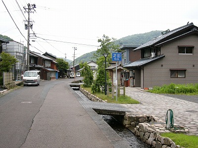 四角く青い「疋田」とかかれた看板と運河について書かれた案内板を旧道、石畳の道とともに遠くから撮影。