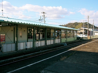 左に薄緑の壁と水色の屋根の平屋の駅舎。