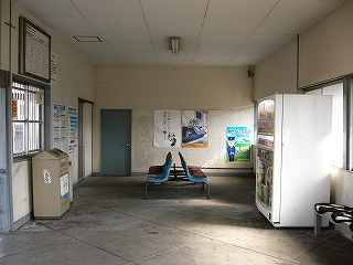 駅舎内を縦長に見て。中央に背中合わせの椅子が置かれている。右手の壁沿いにも椅子、そして自販機。